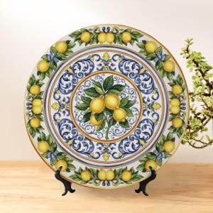 May include: A decorative ceramic plate featuring a lemon design. The plate has a white background with a blue and yellow floral pattern and a gold rim. The center of the plate showcases a cluster of lemons with green leaves.