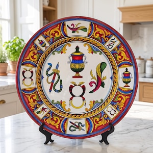 May include: A round ceramic decorative plate featuring a white center with colorful illustrations of a central urn, a snake-wrapped sword, a horn, and a symmetrical emblem. The border displays red, blue, and yellow ornate scrollwork patterns.
