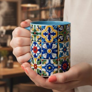 Op de afbeelding: Een keramische mok met een blauw handvat en binnenkant. De buitenkant heeft een kleurrijk, patroon van bloemen tegels in blauw, geel, rood en groen. Stoom komt uit de mok, wat duidt op een warme drank.