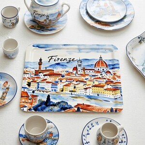 May include: A collection of ceramic tableware featuring a watercolor illustration of Florence, Italy. The set includes a rectangular tray, plates, cups, and a teapot, all with a blue and white color scheme. The word "Firenze" is written on the tray and teapot.