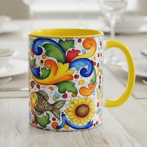May include: A ceramic mug with a yellow handle and interior. The exterior features a colorful floral and fish design with blue, green, orange, and yellow accents. The mug is set on a light-colored wooden table.