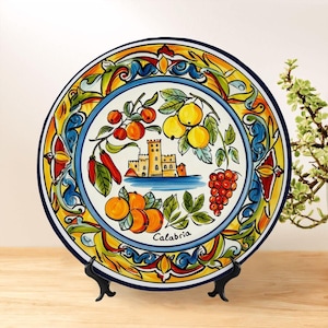 May include: A decorative ceramic plate featuring a colorful design with fruits, a castle, and the word "Calabria." The plate has a blue rim and is displayed on a black stand. The artwork includes oranges, grapes, and lemons.