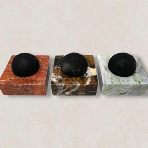 Può includere: Tre oggetti decorativi in pietra. Ognuno presenta una pietra scura e sferica su una base rettangolare. Le basi sono in pietra rossa, marrone e verde. Gli oggetti sono esposti su uno sfondo neutro.