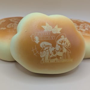 May include: Three flower-shaped buns with a light brown and cream color gradient. The center bun features a cartoon graphic and the text "Battle Subway". The buns are arranged in a close-up shot.