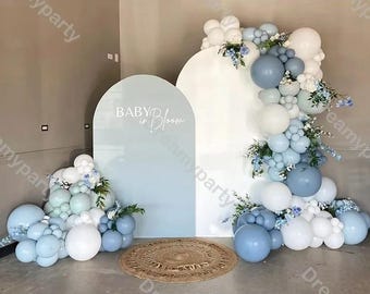 May include: A light blue and white balloon arch with floral accents decorates a baby shower backdrop. The backdrop features two arched panels, one with the text "BABY in Bloom". A woven rug sits in front.