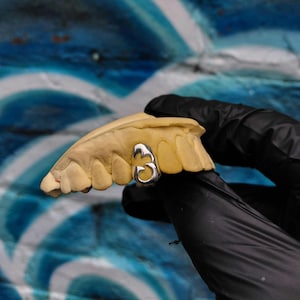 May include: A silver number 3 tooth grill is displayed on a yellow dental mold. The number 3 is in a cursive font. The background is a blue and white painted wall.