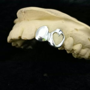 May include: A polished silver dental crown featuring a heart-shaped design, placed on a beige dental mold. The crown's reflective surface contrasts with the dark backdrop. The heart detail provides a decorative touch to the dental appliance.