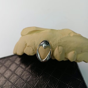 May include: A silver dental tooth cap with a heart-shaped design. The cap is displayed on a beige dental mold, set against a dark, textured surface. The design is simple and elegant.