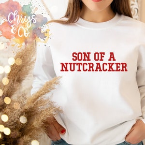 May include: White sweatshirt with red text that reads "Son of a Nutcracker".