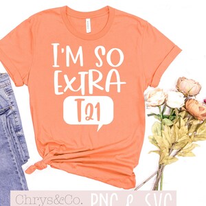 May include: A peach colored t-shirt with the text "I'm so extra T21" in white. The shirt is laid flat on a white surface with a pair of blue jeans and a bouquet of flowers.