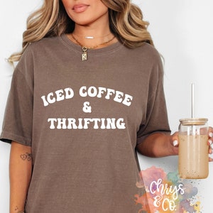 Op de afbeelding: Een bruin t-shirt met de woorden "ICED COFFEE & THRIFTING" in witte, retro-stijl letters. De persoon draagt het shirt en houdt een glas ijskoffie met een rietje vast. Het shirt maakt deel uit van een kledingcollectie.