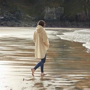 May include: A person wearing a cream-colored hooded fleece jacket walks on a sandy beach with the ocean waves in the background.