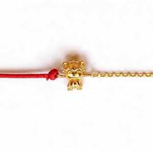 Op de afbeelding: Een goudkleurige bedelarmband met een kleine, gedetailleerde tijgerhanger. De armband heeft aan de ene kant een rood koord, geknoopt, en aan de andere kant een fijne gouden ketting. De achtergrond is wit.