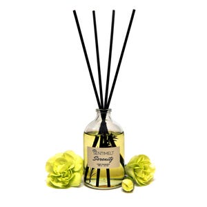 Pode incluir: Um difusor de aroma de vidro com varetas pretas e um rótulo castanho que diz "SENTIMELT Serenity Home Fragrance 100 ml - 3.3 oz". O frasco está cheio de um líquido amarelo e é rodeado por dois cravos verdes.