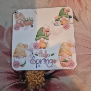 May include: A square, white decorative item featuring watercolor illustrations of gnomes with floral accents. The phrase "Hello Spring" is written in cursive. The gnomes are wearing hats and holding gardening tools.