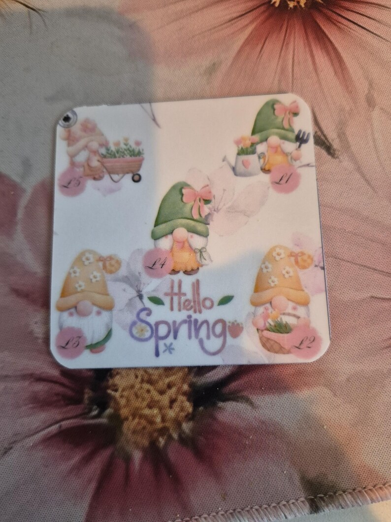 May include: A square, white decorative item featuring watercolor illustrations of gnomes with floral accents. The phrase "Hello Spring" is written in cursive. The gnomes are wearing hats and holding gardening tools.