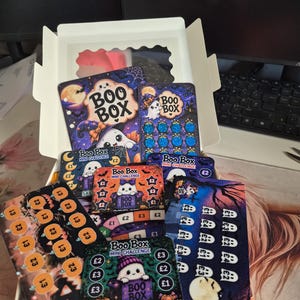 Può includere: Una scatola di cartone bianca contenente diverse schede sfida gratta e vinci Boo Box a tema Halloween. Le carte presentano fantasmi, zucche e pipistrelli con vari valori in sterline, £1, £2, £3 e £4, e la scritta Boo Box.