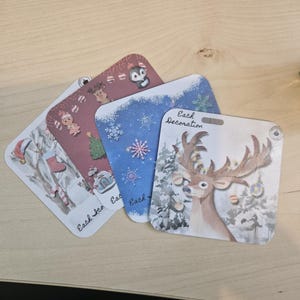 May include: A selection of Christmas gift tags, each with a festive design. The tags feature images of a reindeer, snowman, and penguin. They are square with rounded corners and have the words "Each Decoration" printed on them.
