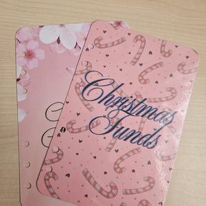 May include: Two pink planner inserts. One has white cherry blossoms, the other a candy cane pattern and the words "Christmas Funds" in a script font. Both have three ring binder holes.