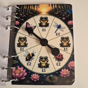 May include: A ring-bound notebook page featuring a circular spinner game with a pond theme. The spinner is divided into sections displaying illustrations of frogs, a butterfly, a dragonfly, a bee, and a ladybug, each labelled with £1 or £2.