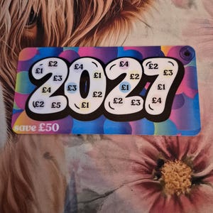 May include: A colourful card featuring the year "2027" in large, bubble-style numbers. The card displays various amounts in British pounds, with the text "save £50". The background has a mix of colours and patterns.