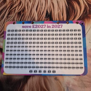 May include: A savings tracker card with the text "save £2027 in 2027". The card is filled with rows of amounts in £5, £1, £10, and £20 increments. The background features a close-up of a Highland cow.