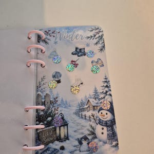 May include: A ring-bound planner page featuring a winter scene with a snowman, a lantern with the text Let it Snow, and various seasonal icons like boots and mittens. The page is decorated with several small, circular, iridescent holographic stickers.