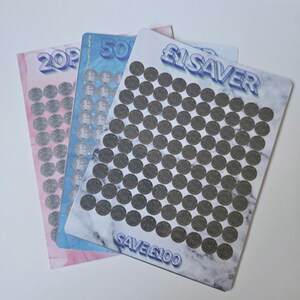May include: Three savings tracker cards in pink, blue, and marble designs. The cards are for 20p, 50p, and £1 savers. The £1 card reads "£1 SAVER SAVE £100". Each card has circles to track savings, ideal for saving money.