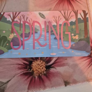May include: A rectangular card with the word "SPRING" in pink, set against a backdrop of a river scene with trees and a blue sky. The card is placed on a floral patterned fabric with pink flowers.