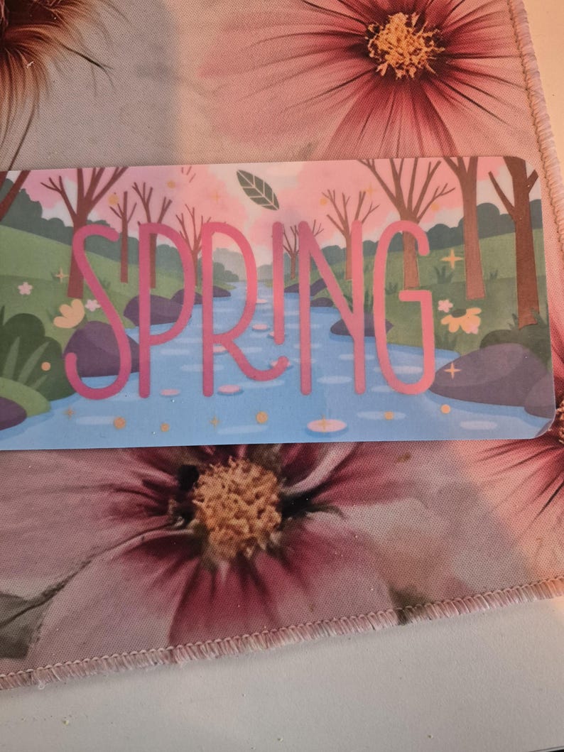 May include: A rectangular card with the word "SPRING" in pink, set against a backdrop of a river scene with trees and a blue sky. The card is placed on a floral patterned fabric with pink flowers.