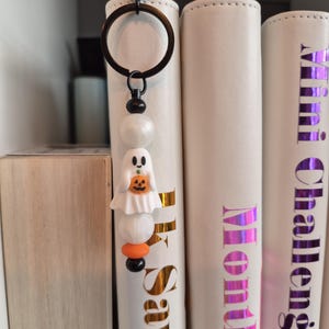May include: A Halloween keyring with a white ghost holding a pumpkin, with white and orange beads. The keyring has a black ring and is in front of white binders with gold and purple text.
