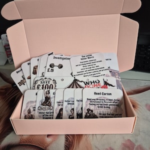 May include: A light pink cardboard box filled with a collection of white game cards featuring black text and illustrations. The cards include character profiles like Victor Norris and Reed Carson, investigation notes, and circus-themed graphics.
