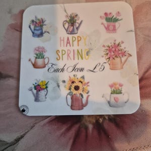 May include: A square sticker featuring watercolor illustrations of watering cans filled with flowers. The text "HAPPY SPRING" is displayed in colorful letters. Each icon is priced at &pound;5.