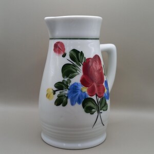Op de afbeelding: Witte keramische kan met een handvat, versierd met handgeschilderde bloemen in rood, blauw en geel. De kan heeft een groene rand aan de bovenkant en een geribbelde basis. Een traditioneel item voor het serveren van drankjes.