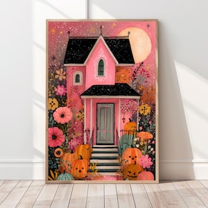 Op de afbeelding: Een ingelijste illustratie van een roze huis met een zwart dak, omringd door bloemen en pompoenen. Een volle maan staat op de achtergrond. Het huis heeft een lichtblauwe deur en trappen naar de veranda. Het kunstwerk heeft een grillig Halloween-thema.