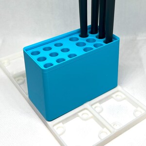 May include: A blue plastic paintbrush holder with 18 holes, holding four paintbrushes. The holder is sitting on a white plastic tray with two compartments.