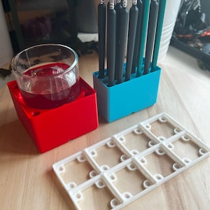 Puede incluir: Un soporte cuadrado rojo con un vaso transparente y líquido rojo, un soporte rectangular azul con pinceles y un objeto blanco en forma de rejilla están sobre una superficie de madera. Los pinceles tienen mangos negros y cerdas transparentes.