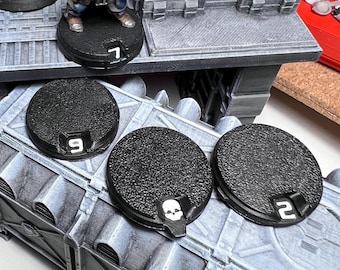 May include: Three black plastic bases with numbers 9, 2, and a skull design. The bases are on a gray and white textured surface. Two blue and yellow painted miniature figures are standing on the bases.