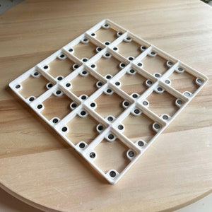 Peut inclure: Grille carrée en plastique blanc avec 25 petits cercles, chacun contenant un point noir. La grille est sur une surface en bois brun clair.