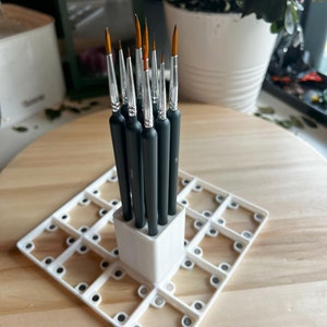 Puede incluir: Un soporte para pinceles de plástico blanco con una base cuadrada y un soporte rectangular. El soporte tiene 16 ranuras para pinceles y está lleno de 10 pinceles negros y plateados.