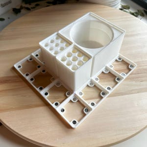 Puede incluir: Organizador de escritorio de pl&aacute;stico blanco con m&uacute;ltiples compartimentos, incluyendo una taza redonda, y una base cuadrada con cierres magn&eacute;ticos.