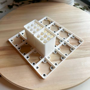 Puede incluir: Organizador de pl&aacute;stico blanco con m&uacute;ltiples compartimentos para guardar objetos peque&ntilde;os. El organizador est&aacute; formado por dos secciones apilables con agujeros circulares y una base con un patr&oacute;n de cuadr&iacute;cula.
