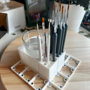 Puede incluir: Portacepillos de pintura de pl&aacute;stico blanco con m&uacute;ltiples ranuras para cepillos. El soporte tiene un vaso de vidrio transparente para agua y una base con m&uacute;ltiples ranuras para otros suministros. El soporte est&aacute; lleno de una variedad de pinceles.