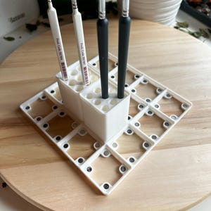 Puede incluir: Portacepillos de pintura de pl&aacute;stico blanco con una cuadr&iacute;cula de compartimentos. Cuatro pinceles est&aacute;n insertados en el portacepillos. El portacepillos est&aacute; sobre una superficie de madera.