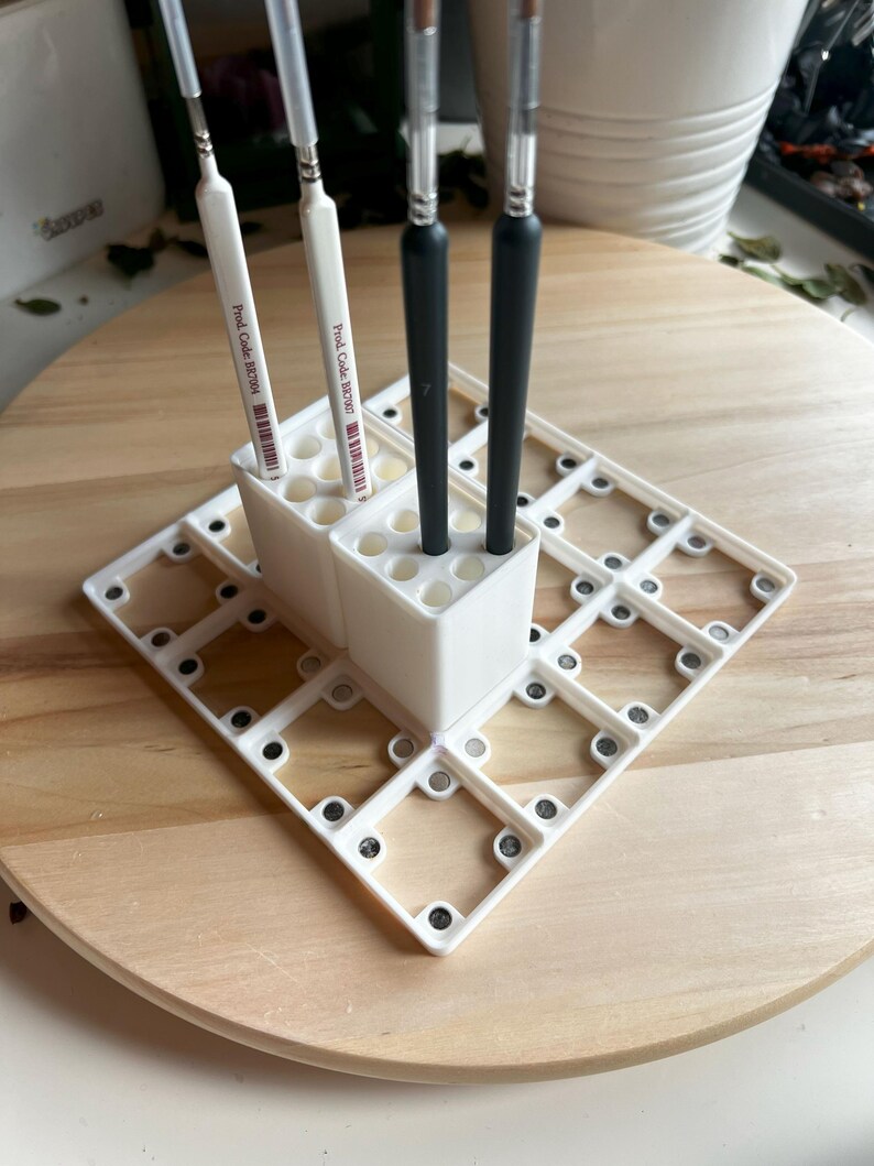 Puede incluir: Portacepillos de pintura de pl&aacute;stico blanco con una cuadr&iacute;cula de compartimentos. Cuatro pinceles est&aacute;n insertados en el portacepillos. El portacepillos est&aacute; sobre una superficie de madera.