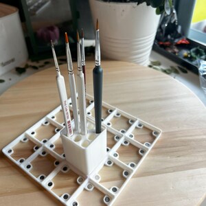 Puede incluir: Portacepillos de pintura de pl&aacute;stico blanco con una cuadr&iacute;cula de compartimentos y un compartimento separado para sostener los cepillos en posici&oacute;n vertical. Seis pinceles est&aacute;n en el portacepillos, incluido un pincel negro con un mango de madera.