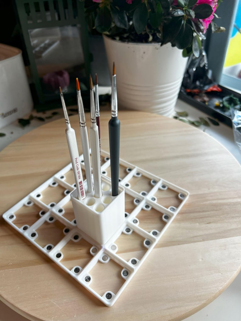 Puede incluir: Portacepillos de pintura de pl&aacute;stico blanco con una cuadr&iacute;cula de compartimentos y un compartimento separado para sostener los cepillos en posici&oacute;n vertical. Seis pinceles est&aacute;n en el portacepillos, incluido un pincel negro con un mango de madera.
