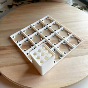 Puede incluir: Rejilla de pl&aacute;stico blanco con peque&ntilde;os c&iacute;rculos negros y un cubo de pl&aacute;stico blanco con nueve agujeros. La rejilla y el cubo est&aacute;n sobre una superficie de madera.