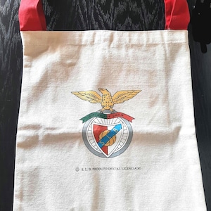 May include: A beige tote bag with red handles. The bag displays a colourful emblem with an eagle, shield, and text. The text at the bottom reads "© S.L.B. PRODUTO OFICIAL LICENCIADO."