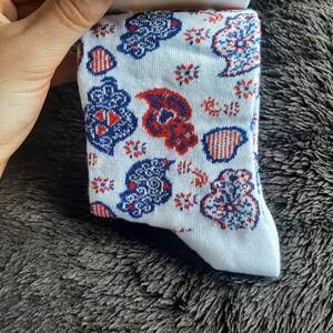 May include: White socks with a repeating pattern of red, white, and blue paisley and floral designs. The socks are held by a hand and are displayed against a dark, textured background.